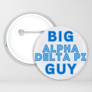 May include: A white button with a blue outline reads "BIG ALPHA DELTA PI GUY". The button has a pin back.