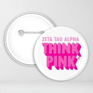 May include: White button with a pink "Think Pink" design and the text "Zeta Tau Alpha" above it. The button has a pin back.