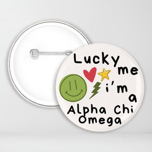 May include: White button with a green smiley face, a red heart, a yellow star, a lightning bolt, and the text "Lucky me I'm a Alpha Chi Omega".