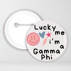 May include: White button with a pink smiley face, a blue heart, a pink star, a white lightning bolt, and the text "Lucky me I'm a Gamma Phi" in black.