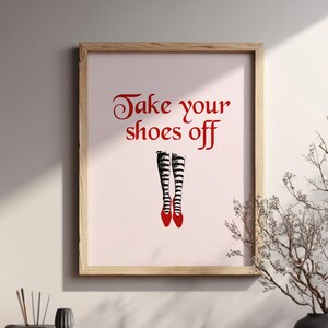Op de afbeelding: Een ingelijste print met de tekst "Take your shoes off" in rode letters boven een illustratie van gestreepte kousen en rode schoenen. Het kunstwerk wordt tentoongesteld op een houten plank met decoratieve items.