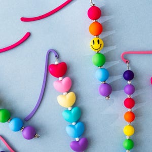 May include: A set of colorful beaded necklaces with heart and smiley face charms. The necklaces are made of bright colors like pink, yellow, blue, green, and purple.