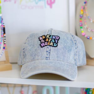 May include: Light blue denim baseball cap with the text "BE KIND & STAY WEIRD" in a colorful patch. The hat is displayed on a white shelf, with other colorful accessories in the background.