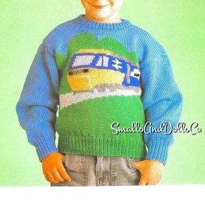 Peut inclure: Un pull en tricot bleu, vert et jaune avec un motif de train de dessin animé. Le pull a un col rond et des manches longues. Le texte "Smalls and Dolls Co." est imprimé en bas du pull.