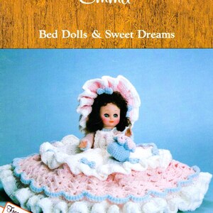 May include: A crocheted doll with a pink and white dress, a blue and white hat, and a blue and white crocheted purse. The doll is sitting on a pink and white crocheted bed. The text "Emma Bed Dolls & Sweet Dreams" is at the top of the image.
