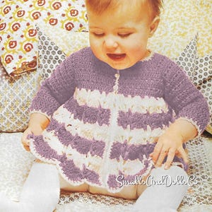 May include: A baby wearing a purple and white crochet dress with long sleeves and a button closure. The dress has a scalloped hem and is paired with white socks. The image is a vintage style photograph.
