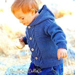 Puede incluir: Un cárdigan azul de punto con capucha, con un patrón de trenza y cierre de botones. El cárdigan lo lleva puesto un niño de pie sobre un tronco.