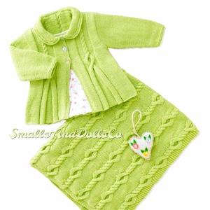 Könnte beinhalten: Eine grüne gestrickte Babyjacke, eine passende Decke und ein kleines herzförmiges Ornament mit Blumenstickerei. Die Jacke hat einen Kragen und einen Knopfverschluss. Die Decke hat ein Zopfmuster.