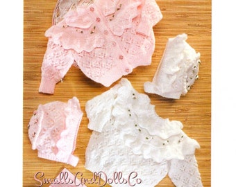 May include: Two knitted baby outfits, one pink and one white, with matching hats. Both outfits feature a ruffled neckline and delicate lace detailing. The outfits are laid out on a wooden surface with the text "Small Girl Dolls Co." visible.
