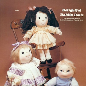 May include: Three handmade fabric dolls, two girls and one boy, with embroidered faces and detailed clothing. The girl dolls are wearing dresses, one with a purple and white checkered pattern and the other with a yellow dress. The boy doll is wearing blue overalls. The dolls are sitting on a wooden chair and a rug with flowers.