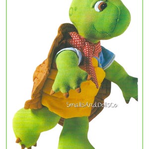 May include: A soft toy turtle with a green head and limbs, a yellow belly, and a brown shell. It is wearing a red polka-dot scarf. The doll has a smiling face and the text "SmallsAndDollsCo".