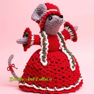 May include: A crocheted mouse doll wearing a red dress with white and green trim, a matching red bonnet, and a long tail with a red bow. The mouse has gray yarn fur, black eyes, and whiskers. The text SmallsAndDollsCo is printed in green and yellow.