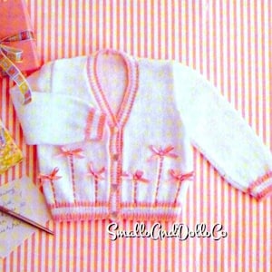 May include: White knitted baby cardigan with pink trim and bows. The cardigan is on a pink and white striped background.