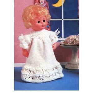 May include: A white knitted doll's dress with a lace trim and a bow on the shoulder. The dress is being worn by a doll with blonde hair and blue eyes.