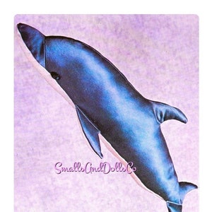 May include: A blue plush dolphin toy with a white belly. The toy is lying on a purple background with the text "Small and Dolls Co." printed in pink.
