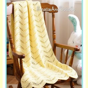 May include: A pale yellow crochet blanket with a chevron pattern draped over a wooden rocking chair. A white plush rabbit toy sits to the right, and a pink and blue plastic rattle rests on the floor. The text SmallsAndDollsCo is visible.