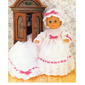Puede incluir: Un vestido de muñeca de bebé de punto blanco con ribete de cinta rosa y un gorro de punto blanco a juego con cinta rosa. La muñeca lleva el vestido y el gorro y está de pie frente a un armario de madera.