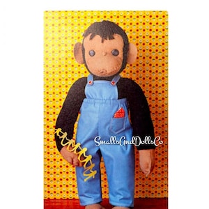 Może przedstawiać: Brązowa i czarna małpka w niebieskich ogrodniczkach z czerwoną chusteczką w kieszeni. Lalka trzyma żółty łańcuch połączonych figurek. Lalka stoi na drewnianej powierzchni z żółtym kwiatowym wzorem w tle. W obrazie widoczny jest tekst "Smalls And Dolls Co".