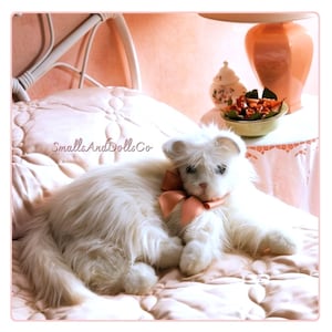 May include: A plush white cat figurine with long, soft-looking fur and a peach-colored satin ribbon tied around its neck. The cat is lying down on a quilted surface. The text SmallsAndDollsCo is visible in the center of the image.