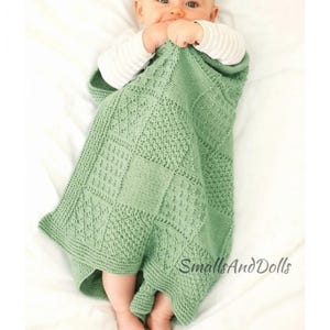May include: A sage green knitted baby blanket featuring a patchwork design with various textured patterns, including cables and seed stitches. The blanket has a border and the text SmallsAndDolls is printed in the lower right corner.