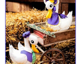 May include: Two white plush goose dolls with yellow beaks and black eyes. One goose wears a purple polka-dot bonnet and apron with lace trim. The other wears a purple plaid hat and a green and white plaid tie. Text reads Smalls Cind Dolls Co.