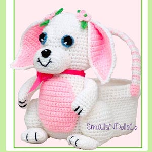 May include: White crocheted dog basket with pink ears, a pink bow, and pink flowers on its head. The basket has a white crocheted handle. The text "SmallIsNDollsCo" is visible on the basket.