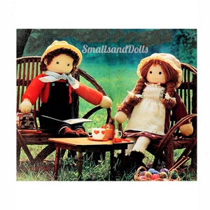 May include: Two knitted dolls sitting at a table with a teacup and saucer. The dolls are wearing overalls and a dress. The dolls are sitting on wicker chairs in a garden setting. The text "SmallsandDolls" is visible in the image.