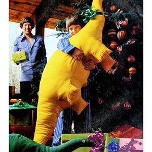 May include: A large, bright yellow, soft-stuffed dinosaur toy with a long neck and tail, featuring stitched details on the feet. The dinosaur is held upright, showcasing its elongated body shape and smooth fabric texture, positioned near a decorated tree.