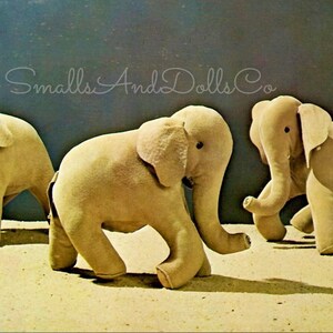 May include: Three plush toy elephants in a light brown color. The elephants are standing on a sandy surface. The elephants have large ears and trunks. The text "Smalls And Dolls Co" is written in white on a blue background.