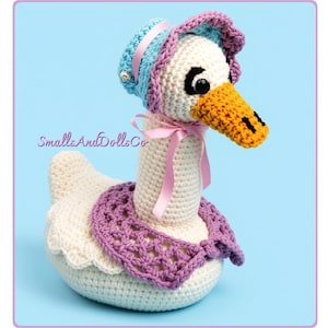 May include: A cream-colored crochet goose doll featuring a yellow beak, black eyes, and a light blue bonnet with a pink ribbon. The goose wears a purple crocheted collar and a pink ribbon bow around its neck. The text SmallsAndDollsCo is visible.
