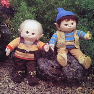 May include: Two plush elf dolls, one with a white beard and brown hair, the other with brown hair and a blue hat. Both dolls are wearing brown pants and colorful shirts. The dolls are sitting on a rock.