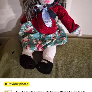 May include: A handmade fabric doll with yarn hair, wearing a blue crocheted hat, a red long-sleeved top with a light blue patterned neck tie, and a teal skirt featuring a pink and white floral print. The doll has black shoes and embroidered facial features.