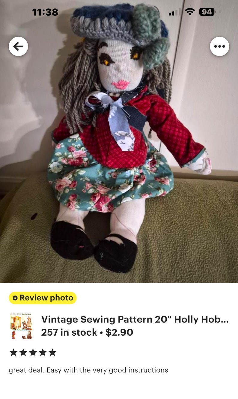 May include: A handmade fabric doll with yarn hair, wearing a blue crocheted hat, a red long-sleeved top with a light blue patterned neck tie, and a teal skirt featuring a pink and white floral print. The doll has black shoes and embroidered facial features.