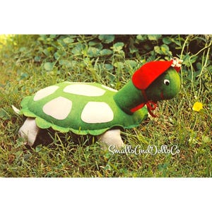Könnte beinhalten: Eine grüne Schildkröte aus Filz mit einem weiß gefleckten Panzer und einer roten Haube, die mit einem Band unter dem Kinn gebunden ist. Die Schildkröte hat ein gesticktes Gesicht mit weißen Augen und schwarzen Pupillen. Der Text SmallsAndDollsCo ist sichtbar.