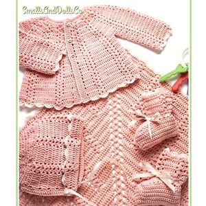 May include: A dusty pink crochet baby set featuring a long-sleeve cardigan with white lace trim, a matching bonnet with ribbon ties, two booties with ribbon bows, and a large blanket. The text SmallsAndDollsCo is visible in the top left corner.