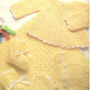 May include: A set of crocheted baby clothes in yellow with white trim. The set includes a cardigan, a bonnet, booties, and a blanket.