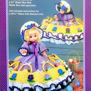 May include: Crochet pattern for a 13-inch music box doll outfit and a 10 1/2-inch pillow doll outfit. The dolls are wearing purple dresses with white lace trim and yellow crocheted baskets with purple flowers. The dolls are sitting on a yellow crocheted base with white lace trim and purple flowers.