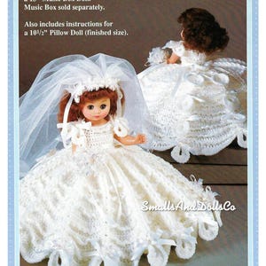 Puede incluir: Un vestido de novia de crochet blanco y un velo para una muñeca de 33 cm. La muñeca lleva el vestido y el velo. El texto "Crochet an outfit to fit a 13" Music Box Doll. Music Box sold separately. Also includes instructions for a 10 1/2" Pillow Doll (finished size)." es visible en la imagen.