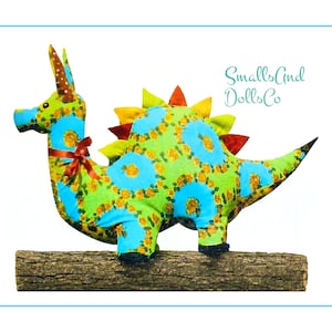May include: A plush dinosaur toy featuring a bright blue and lime green floral pattern, adorned with a brown ribbon bow at the neck and colourful fabric spikes along its back. The text SmallsAndDollsCo is printed in blue on the upper right side.