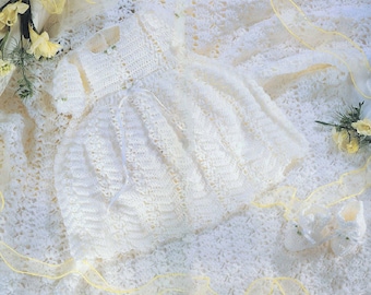 May include: A white crocheted baby dress with a matching bonnet and booties. The dress has a delicate lace pattern and a satin ribbon tie.