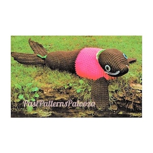 May include: A crocheted brown and pink sea lion plush toy with black eyes and a green nose. The toy is lying on a bed of green grass.