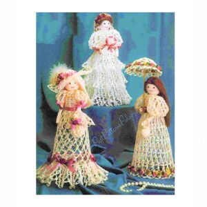 May include: Three crocheted dolls dressed in white and pink dresses with lace trim. The dolls are holding flowers and a parasol. The dolls are standing on a blue background.