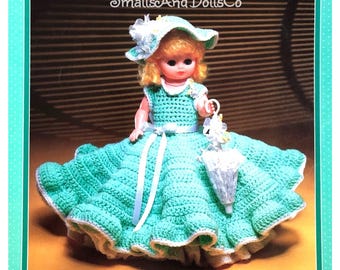 May include: A crocheted doll wearing a green dress with a white sash and a white hat with yellow flowers. The doll is holding a white umbrella with yellow flowers.