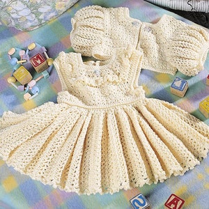 May include: A crocheted baby dress and bonnet set in a cream color. The dress has a ruffled neckline and a gathered skirt. The bonnet has a simple design.