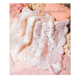 May include: A white crocheted baby blanket with a scalloped edge and a white satin ribbon tied around it. The blanket is on a pink and white floral patterned surface.