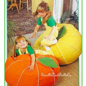 May include: Two children in green and yellow outfits play on large, orange and yellow beanbag chairs shaped like fruit. The orange beanbag has a green leaf and white stitching. The yellow beanbag also has a green leaf.
