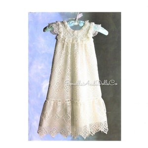 Puede incluir: Un vestido de bebé blanco de ganchillo con un dobladillo festoneado y mangas con volantes. El vestido está colgado de una percha azul y tiene el texto "Small Arts Dolls Co." en la parte inferior.