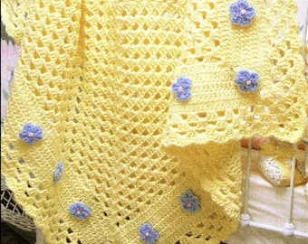 May include: A yellow crocheted baby blanket with a crocheted border and purple flowers. The blanket is on a white cot.