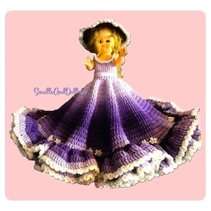Peut inclure: Une poupée vêtue d'une robe crochetée avec un dégradé violet, dotée d'une jupe large avec une bordure blanche à volants et de petites décorations florales. La poupée porte un chapeau assorti à larges bords. Le texte SmallsAndDollsCo est visible.