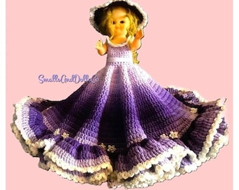 May include: A doll wearing a crocheted dress with a purple ombre gradient, featuring a wide skirt with ruffled white trim and small flower embellishments. The doll wears a matching wide-brimmed hat. The text SmallsAndDollsCo is visible.